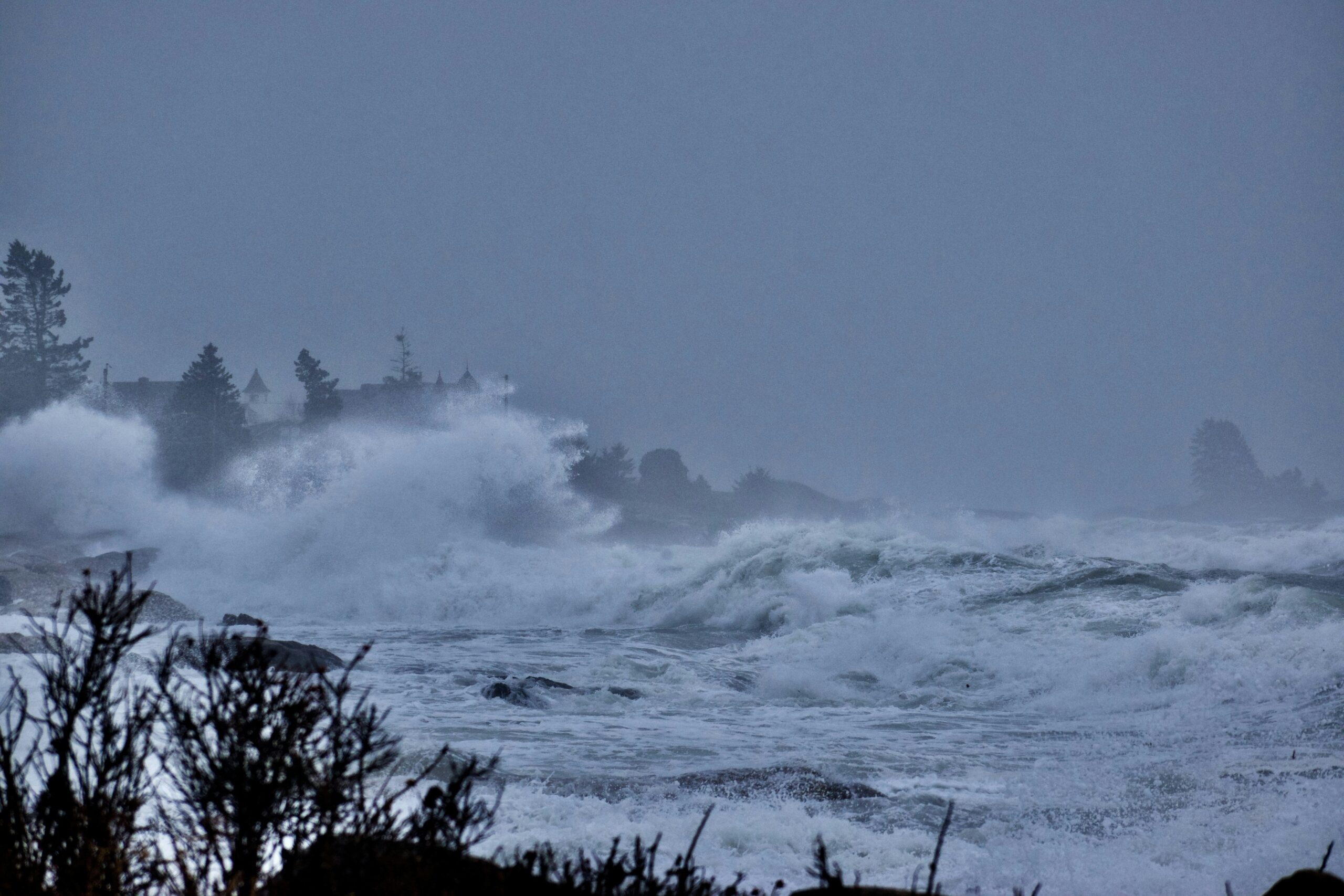 Dark, overcast sky over a stormy coastline with huge waves crashing toward the shore, distant trees and buildings barely visible through the mist and spray, evoking the destructive power and chaos of a hurricane.